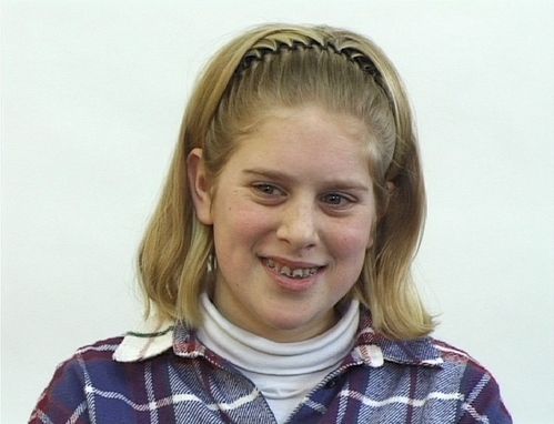 A blonde girl with a headband and braces smiles and looks slightly embarrassed to the side