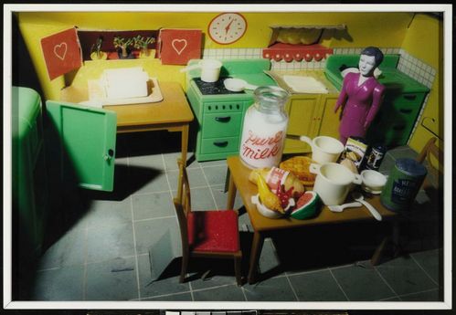 The yellow-painted kitchen of a dollhouse is photographed from an elevated, oblique angle. In the corner of the kitchen stands a doll figure wearing a glossy purple dress. Various foods are spread across the table. Particularly prominent is a milk container with the red label “Pure Milk”.