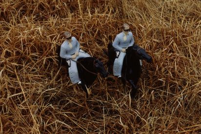 Aus der Vogelperspektive blickt man auf zwei männliche Figuren, die durch ein Ährenfeld reiten. Sie tragen hellblaue Kleidung und sitzen auf schwarzen Pferden. Beide tragen schwarze Banditenmasken mit ausgeschnittenen Augen.
