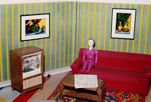 In front of blue-and-yellow striped wallpaper, a doll sits on a red sofa. The living room is furnished in an old-fashioned style, featuring a wood-paneled television and an Oriental rug. Still lifes hang on the walls.