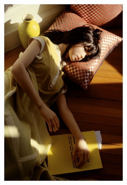 A realistic, life-sized doll figure lies in a lime-green dress on an orange-and-silver checked cushion. Behind it stands a vase in the same lime-green colour. Beneath its slightly outstretched hand lies a yellow exhibition catalogue. The doll stares into nothingness with its empty plastic eyes.