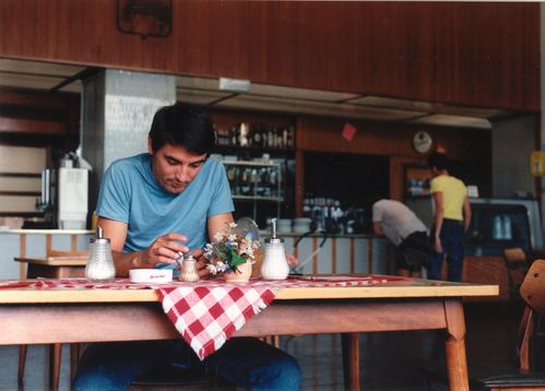 A man in a light blue T-shirt sits at a restaurant table with a red and white checkered tablecloth.
