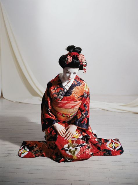 A hyperrealistic, life-sized doll is dressed as a geisha. Her hair is styled into an elaborate bun, woven with red floral ornaments. Her face is painted white, in contrast to her hands. She wears a silk kimono in red and deep blue tones.