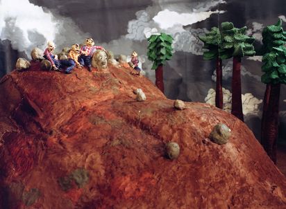 Auf einem rotbraunen Hügel sitzen vier männliche Knetfiguren inmitten von Steinen. Die Männer haben alle karierte Hemde und Latzhosen an. Ein Stein hat ein Gesicht, andere rollen den Hügel hinunter. Hinter dem Hügel stehen hohe Bäume vor einem grauen Wolkenhimmel. 