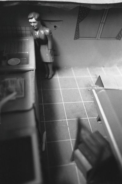 Taken from a slightly elevated angle, the black-and-white portrait-format photograph shows a kitchen interior. On the left side, a kitchen unit is visible, while on the right a table and chair are suggested. The image focuses on a doll figure wearing a suit, who is looking into a sink at the far end of the kitchen counter.