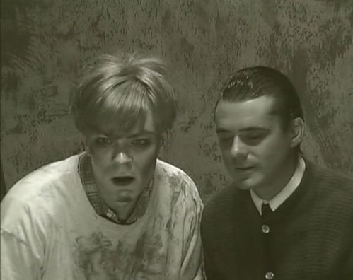 Schwarzweißer Video- Screenshot: Zwei männliche Figuren, die erste mit zotteliger blonden Frisur und schmutzigem T-Shirt, die zweite mit nach hinten gegelten Haaren und Janker