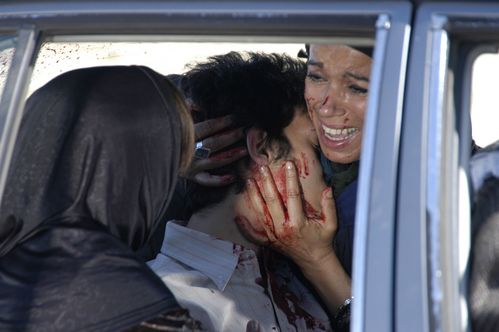 In the back seat of a car, a desperate woman in a headscarf hugs the bloodied head of a man. Another woman with a headscarf sits next to the wounded man with her back to the viewer.