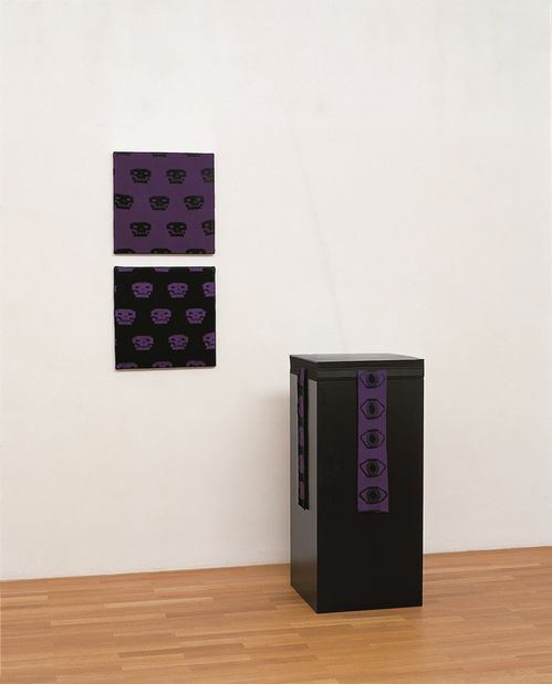 Werkgruppe bestehend aus zwei Strickbildern in Schwarz und Lila an der Wand, die ein Totenkopfmuster zeigen, sowie einem schwarzen quadratischen Sockel mit schwarz-lilafarbenen gestrickten Textilbahnen versehen, die Augenmotive zeigen.