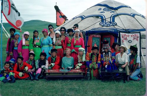 Vor einer mongolischen Jurte steht eine Gruppe von Menschen. Die meisten tragen traditionelle mongolische Kleidung, einige wenige sind in auffälliger europäischer Kleidung gekleidet. Die verschiedenen farbigen Gewänder machen das Bild farbenfroh. Ein kühler Blaustich über der Szene.