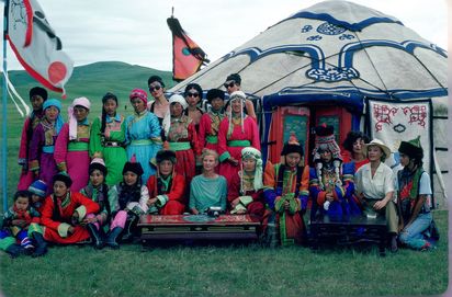 Vor einer mongolischen Jurte steht eine Gruppe von Menschen. Die meisten tragen traditionelle mongolische Kleidung, einige wenige sind in auffälliger europäischer Kleidung gekleidet. Die verschiedenen farbigen Gewänder machen das Bild farbenfroh. Ein kühler Blaustich über der Szene.