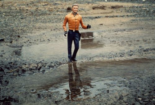 Eine männliche Puppenfigur, die an eine Actionfigur erinnert, läuft durch eine Pfütze. Sein orangefarbenes Oberhemd und ein Holster erinnern an die Kleidung eines Cowboys.