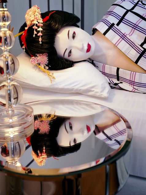 Eine hyperrealistische, lebensgroße Puppe ist als Geisha gekleidet. Ihr Haar ist zu einer aufwendigen Duttfrisur hochgesteckt, in die roter Blumenschmuck eingearbeitet ist. Ihr Gesicht ist weiß geschminkt, und sie trägt einen weißen, karierten Kimono. Ihr Kopf ruht auf einem Kopfkissen. Der danebenstehende spiegelnde Nachttisch reflektiert das Gesicht der lebensgroßen Puppe.