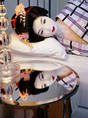 Eine hyperrealistische, lebensgroße Puppe ist als Geisha gekleidet. Ihr Haar ist zu einer aufwendigen Duttfrisur hochgesteckt, in die roter Blumenschmuck eingearbeitet ist. Ihr Gesicht ist weiß geschminkt, und sie trägt einen weißen, karierten Kimono. Ihr Kopf ruht auf einem Kopfkissen. Der danebenstehende spiegelnde Nachttisch reflektiert das Gesicht der lebensgroßen Puppe.