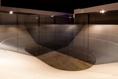 Innenansicht eines Architekturmodells: eine halbkreisförmige Holzstruktur, die an eine Stadiontribüne erinnert, wird von zwei Glaswänden gespiegelt. Daraus entsteht die optische Illusion einer durchgehenden ovalen Architektur.