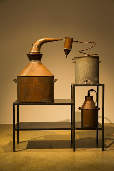 Different sized kettles made of copper and gray metal on a black shelf. Partially they are connected by a spiral tube.