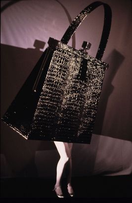 Vor einem lilafarbenen Hintergrund steht auf Frauenbeinen in hellen Strumpfhosen und Pumps eine große schwarze Handtasche in Krokodillederoptik, die den Oberkörper der Figur bildet. Die Konturen der Tasche werfen starke Schatten auf den lilafarbenen Stoffhintergrund.
