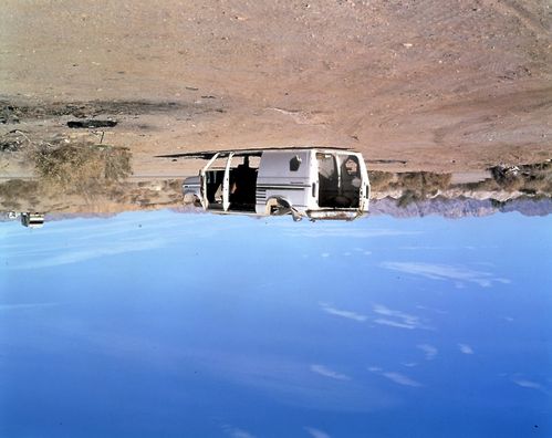 An overturned, burned-out van is shown in a desert landscape beneath a bright blue sky. The image is rotated, making the sky appear to form the ground.