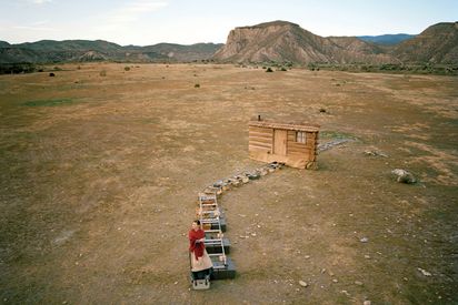 In the middle of a barren desert landscape, like a movie set, there is a short section of railroad tracks and a wooden house wall. At the beginning of the railroad tracks stands a woman with a red scarf over her shoulders.