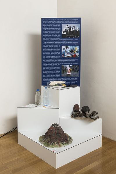 A display with several platforms, with information about the work displayed at the very top. Below that, various objects are arranged in tiers, including bottles and a model of a volcano.