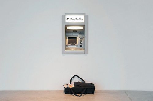 In eine weiße Wand ist ein Geldautomat eingebaut. Darüber befindet sich eine weiße Leuchttafel mit der Aufschrift „24-Hour Banking“. Vor dem Automaten steht eine schwarze Tragetasche, in der ein kleines Kind liegt.