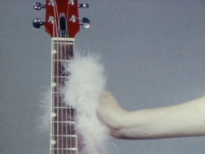 A hand holds the neck of a red guitar with a pale pink fur puff.