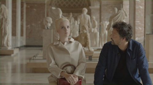 Ein Mann und eine Frau sitzen nebeneinander auf einer Museumsbank, hinter ihnen sind Gipsabdrücke antiker Skulpturen. Der Mann ist hat seinen Kopf zu der Frau zugewandt.
