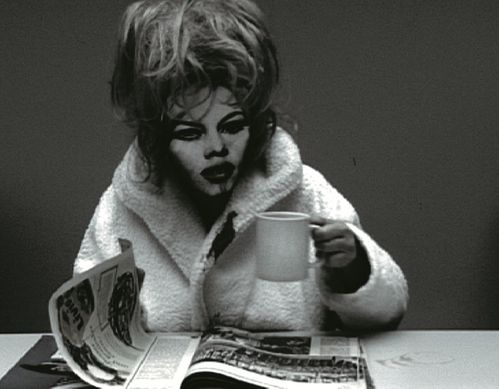 Standbild aus einem Video. Man sieht eine Person in Bademantel, die eine Tasse anhebt und eine Zeitschrift vor sich auf dem Tisch umblättert. Sie hat aufgetürmte blonde Haare und trägt eine Maske mit einem stark geschminkten Gesicht darauf.