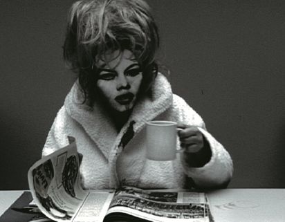 Standbild aus einem Video. Man sieht eine Person in Bademantel, die eine Tasse anhebt und eine Zeitschrift vor sich auf dem Tisch umblättert. Sie hat aufgetürmte blonde Haare und trägt eine Maske mit einem stark geschminkten Gesicht darauf.